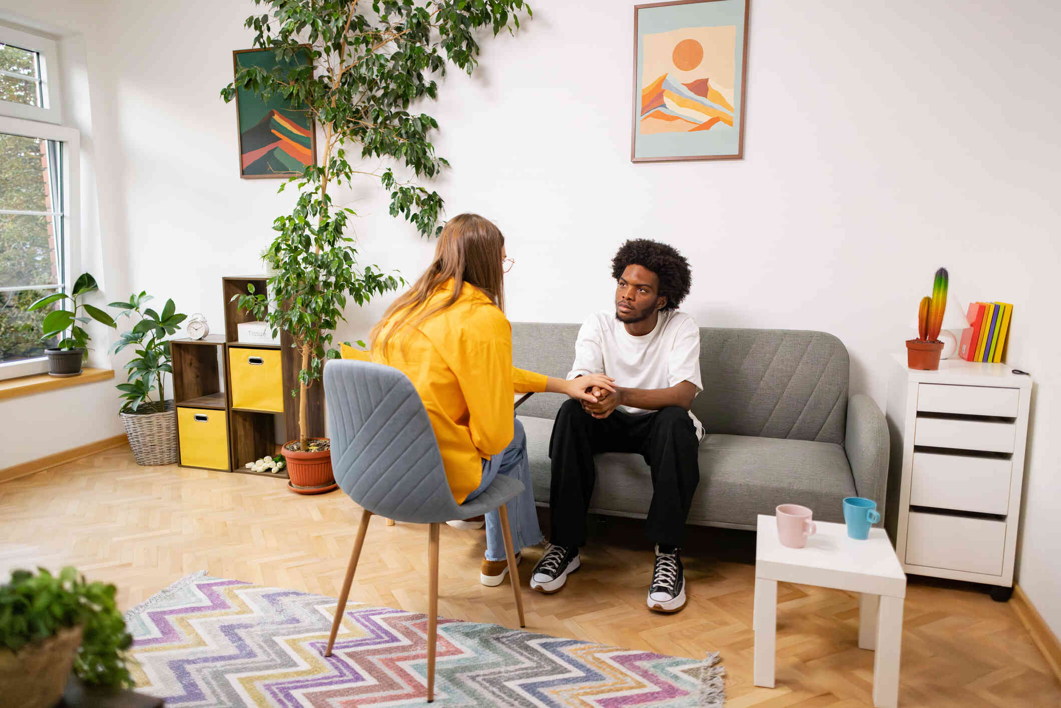 A young man in a white shirt with a neutral facial expression sits on a couch in a therapist office while engaged in a conversation with a woman in a yellow shirt